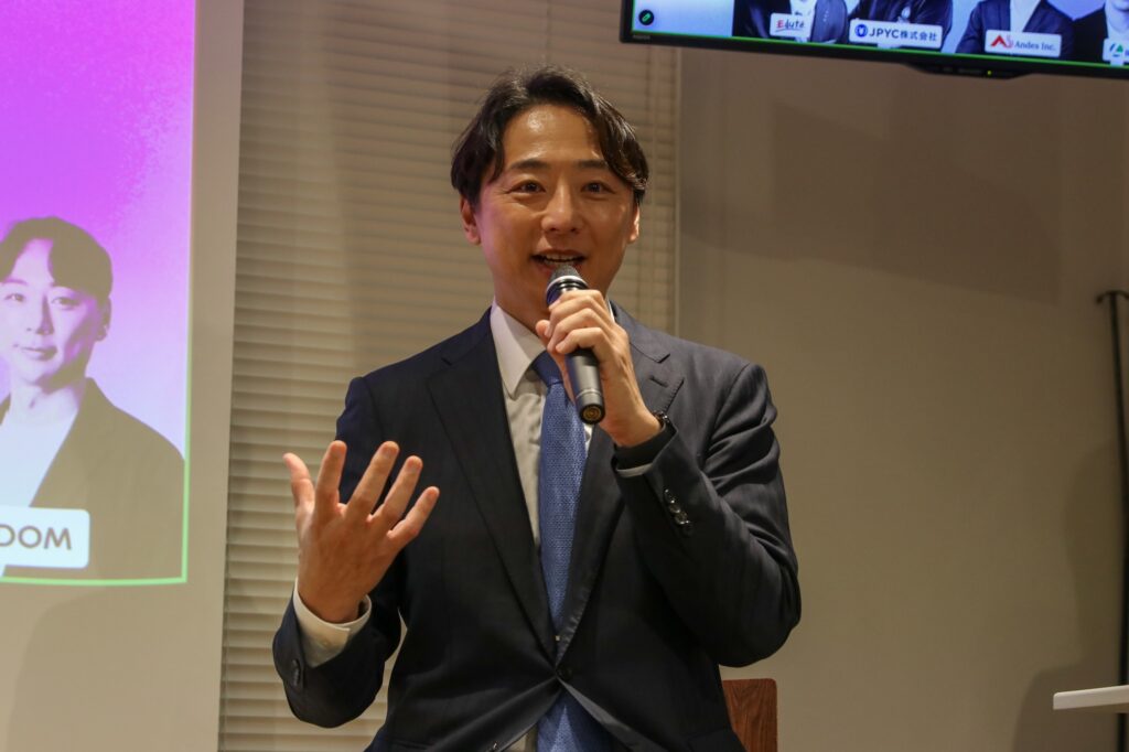Man in a dark suit and blue tie speaks into a handheld microphone, gesturing with his left hand in a conference room.