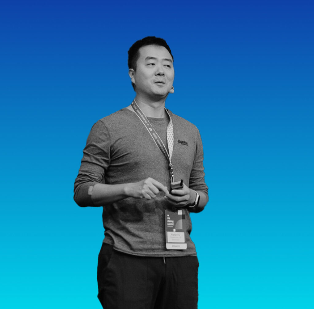 Male conference speaker wearing a lanyard and badge, gesturing while presenting on a blue gradient stage backdrop